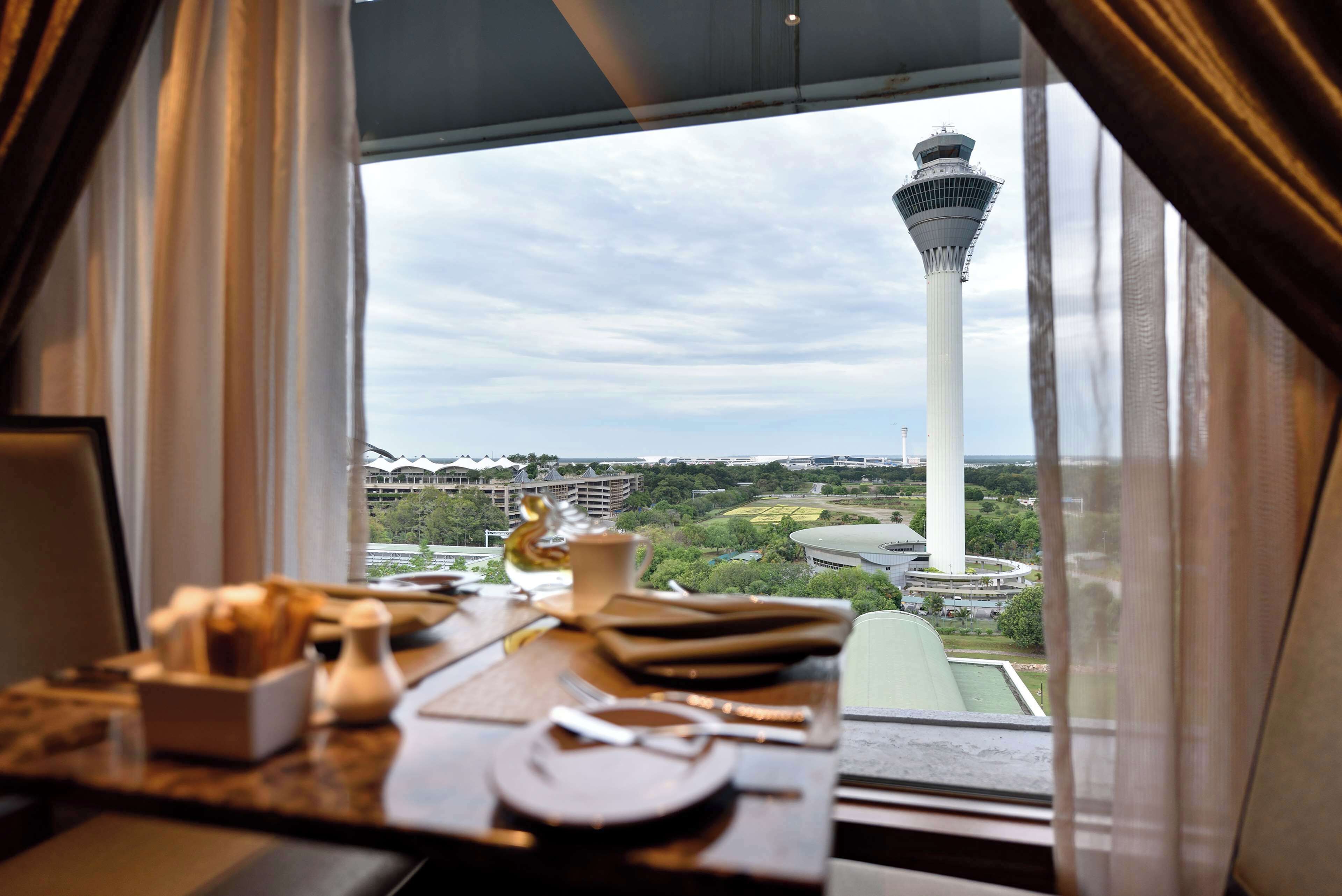 Mesa de comedor con platos, cubiertos y salero, con vistas a una torre y vegetación.