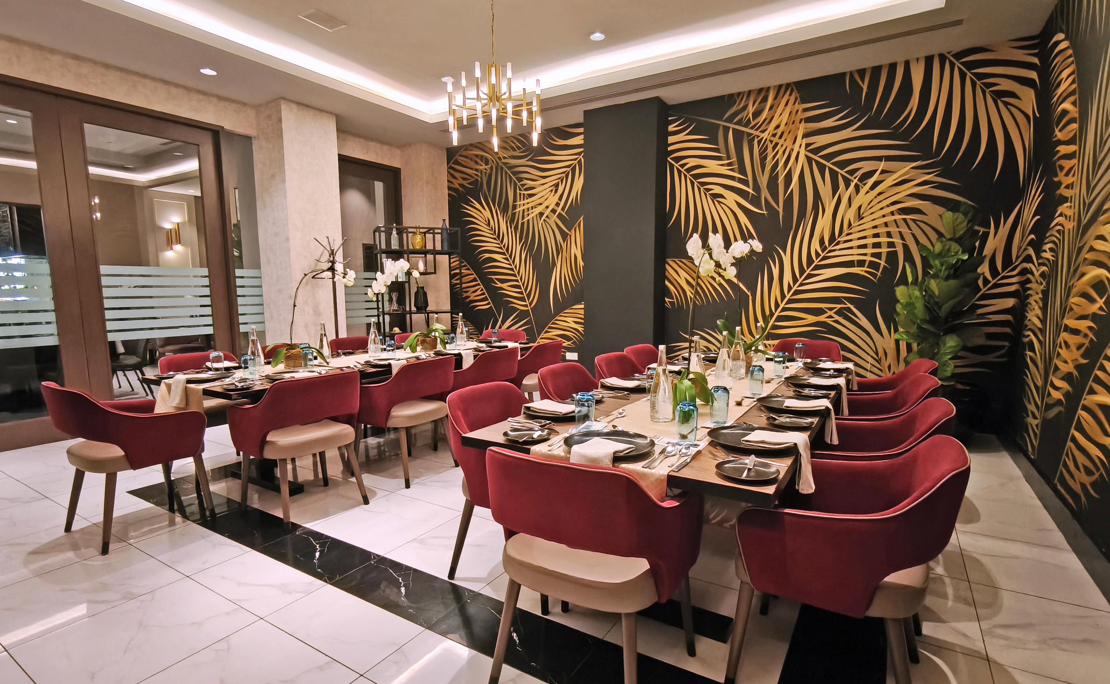 A dining area with red chairs, a long table set for a meal, and a wall with a tropical leaf pattern.