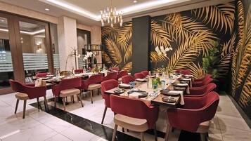 A dining area with red chairs, a long table set for a meal, and a wall with a tropical leaf pattern.