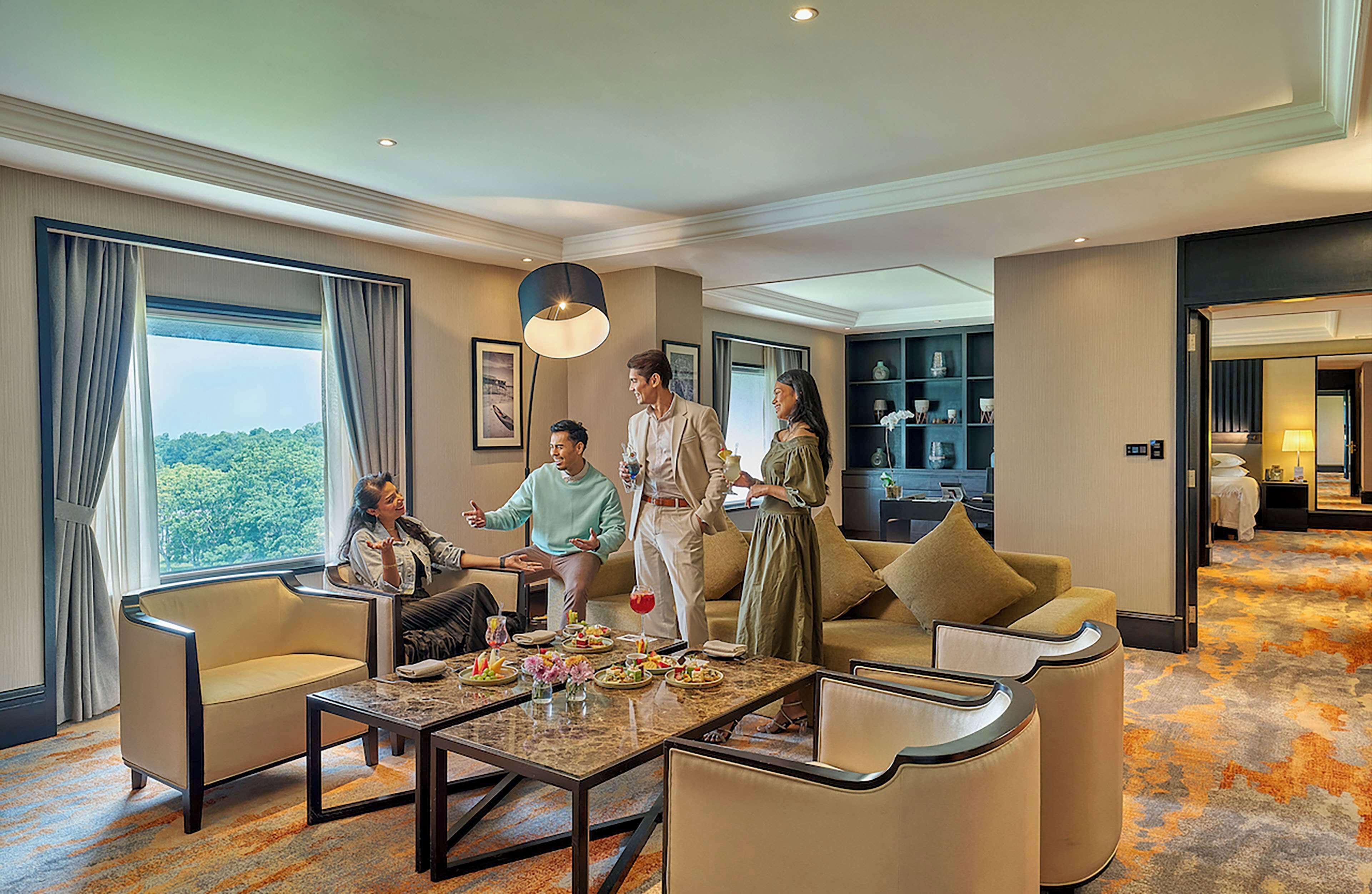 A hotel room with a large window, a sofa, a coffee table, and a dining area with a chandelier.