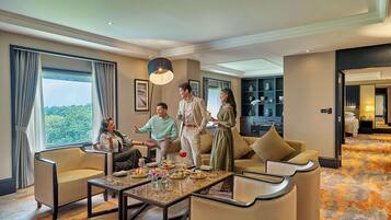 A hotel room with a large window, a sofa, a coffee table, and a dining area with a chandelier.