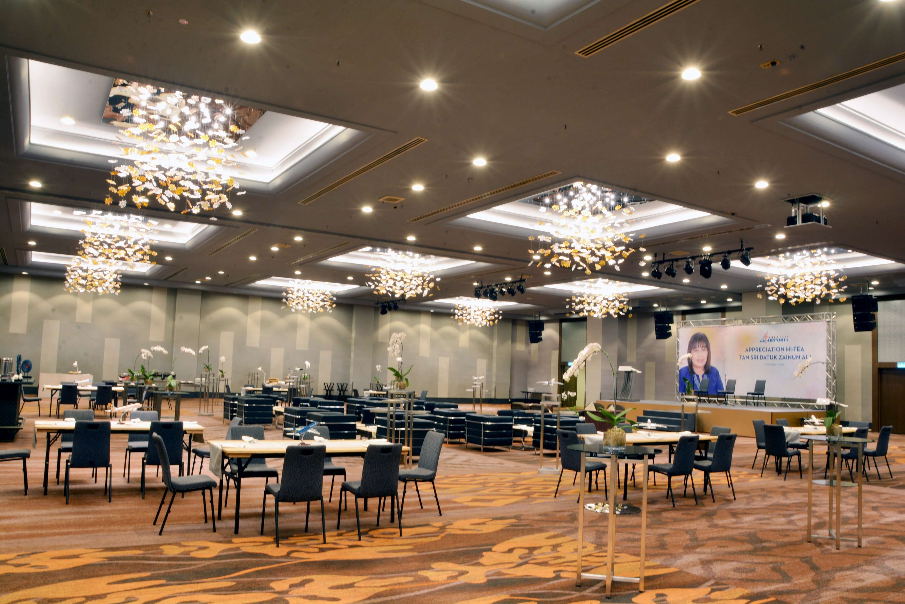 Uma sala de conferências ampla, com várias mesas e cadeiras, um palco com tela de projeção e lustres modernos.