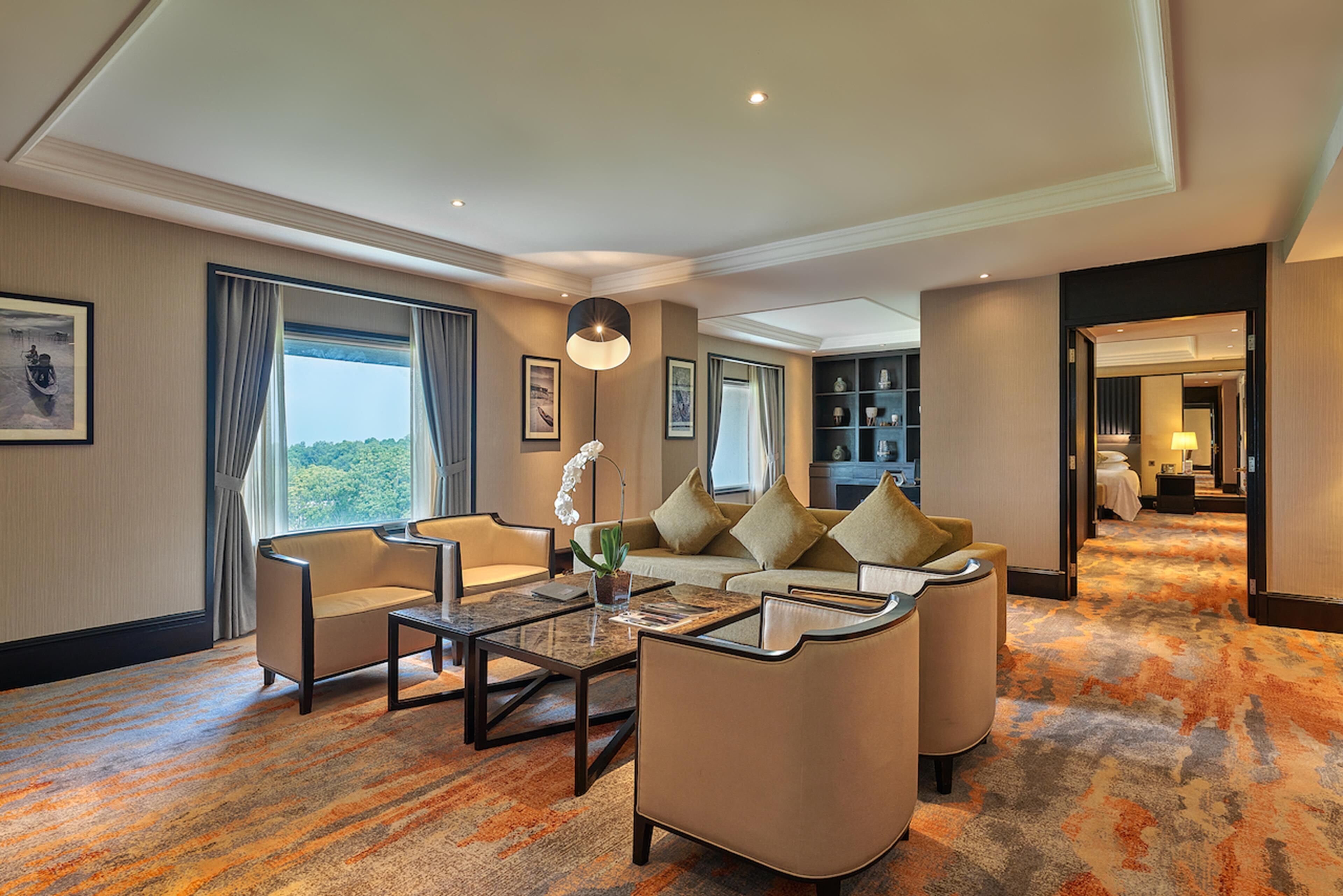 A hotel room with a sofa, armchairs, a coffee table, a large window with curtains, a bookshelf, and a carpeted floor.