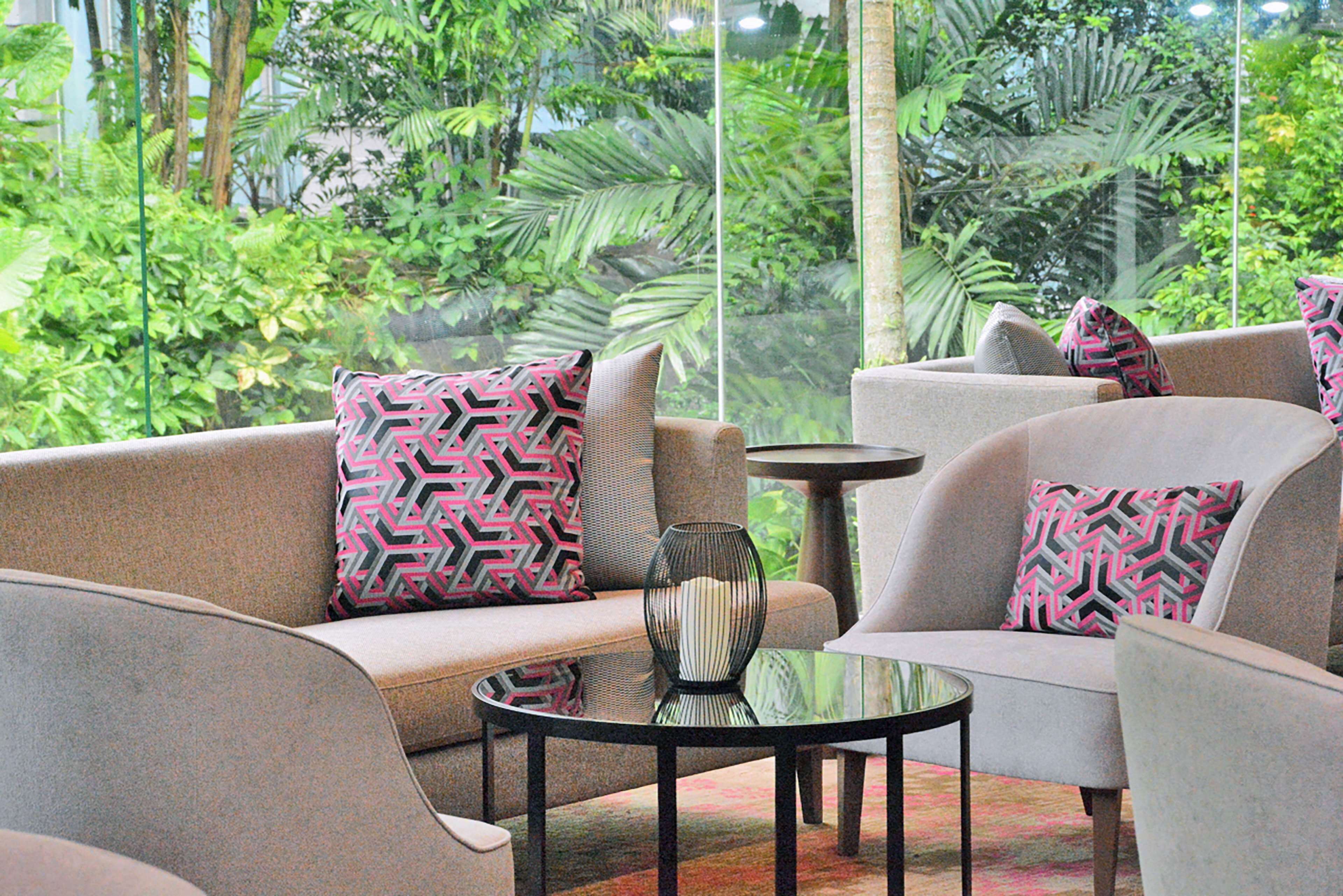 A seating area with sofas and armchairs, a round table, and a view of lush greenery through large windows.