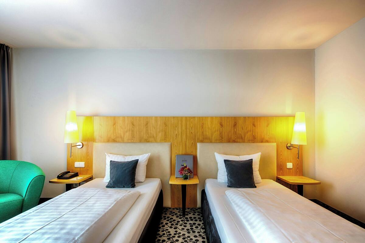 Une chambre d’hôtel avec deux lits, une tête de lit en bois, des lampes de chevet, un téléphone et une petite table sur laquelle est posé un vase de fleurs.