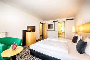 Een hotelkamer met een bed, een groene fauteuil, een tafeltje met een vaas en een spiegel aan de muur.