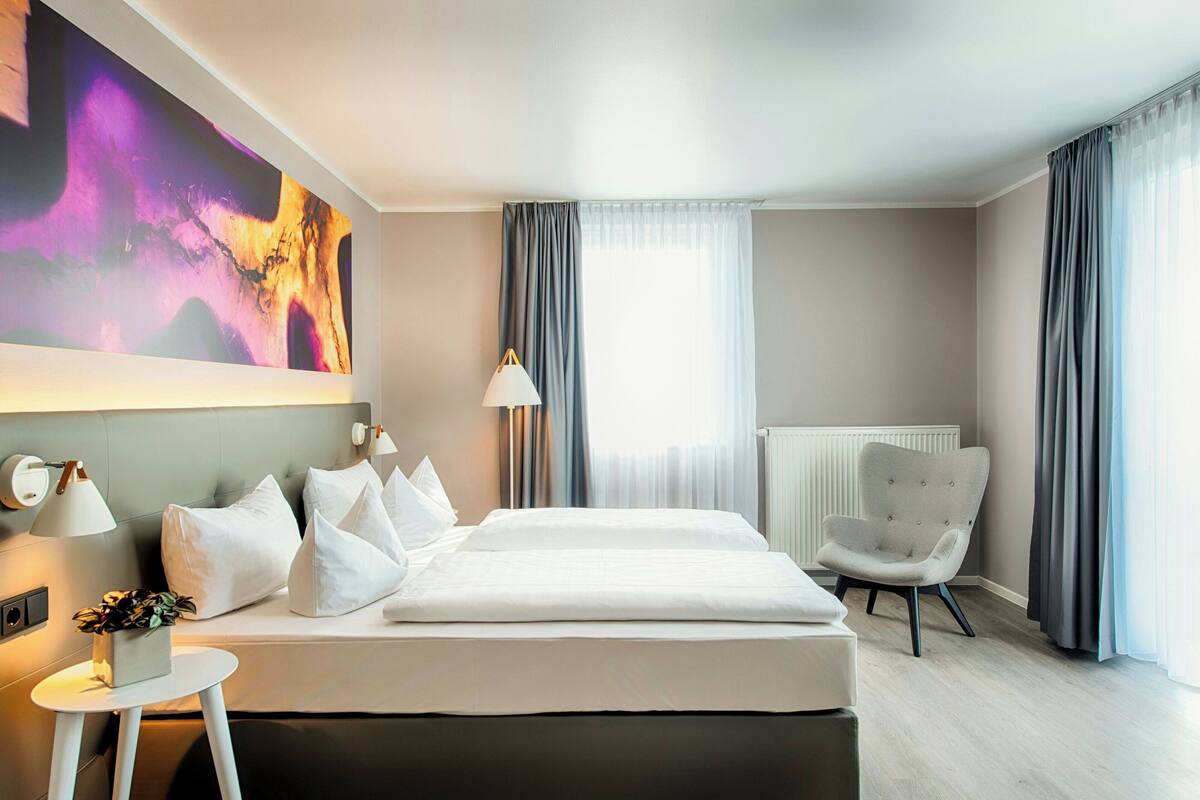 Une chambre d’hôtel moderne avec un grand lit, une chaise, une petite table avec une plante et un grand tableau abstrait accroché au mur.