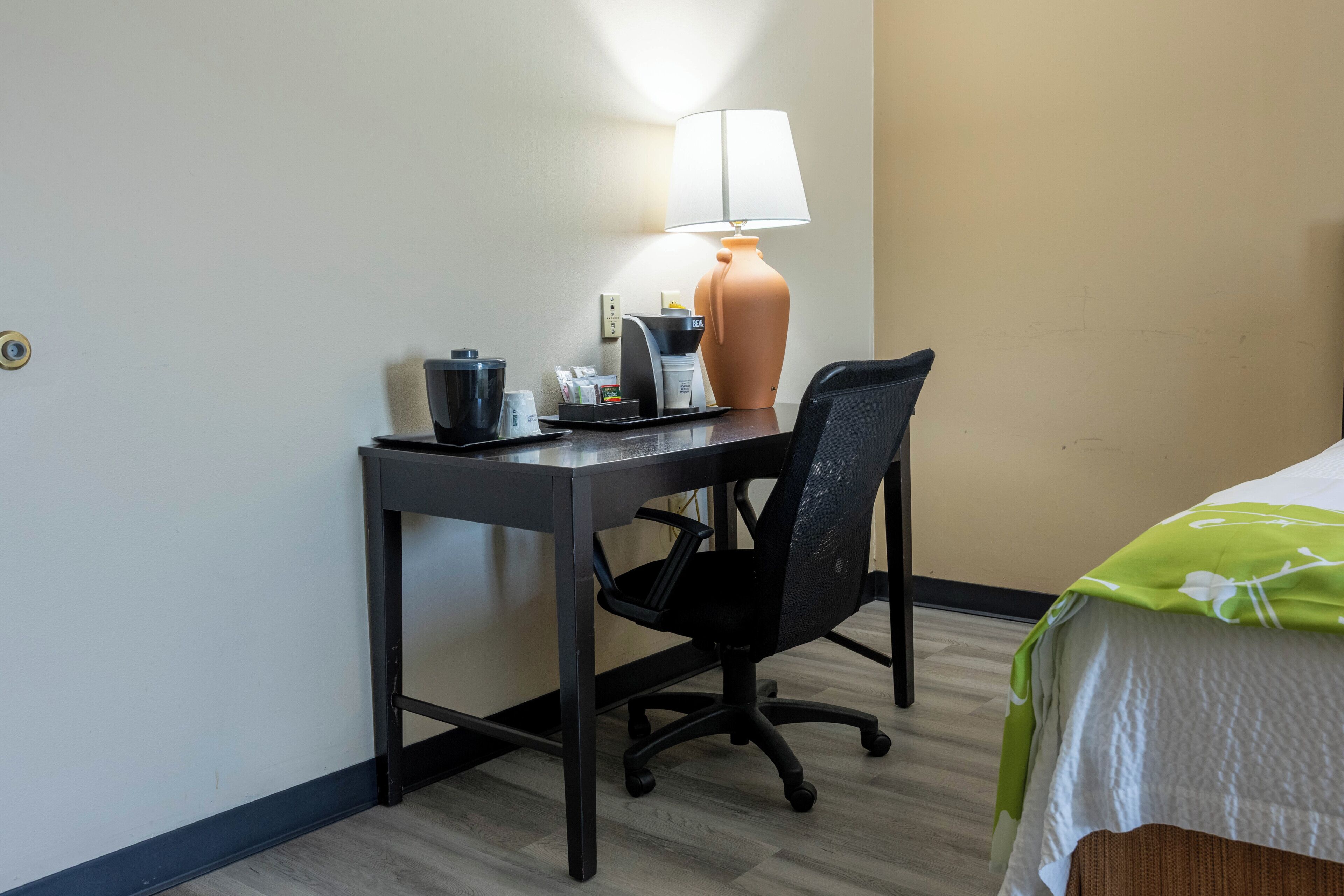 A hotel room with a desk, chair, lamp, and a bed with a green floral bedspread.
