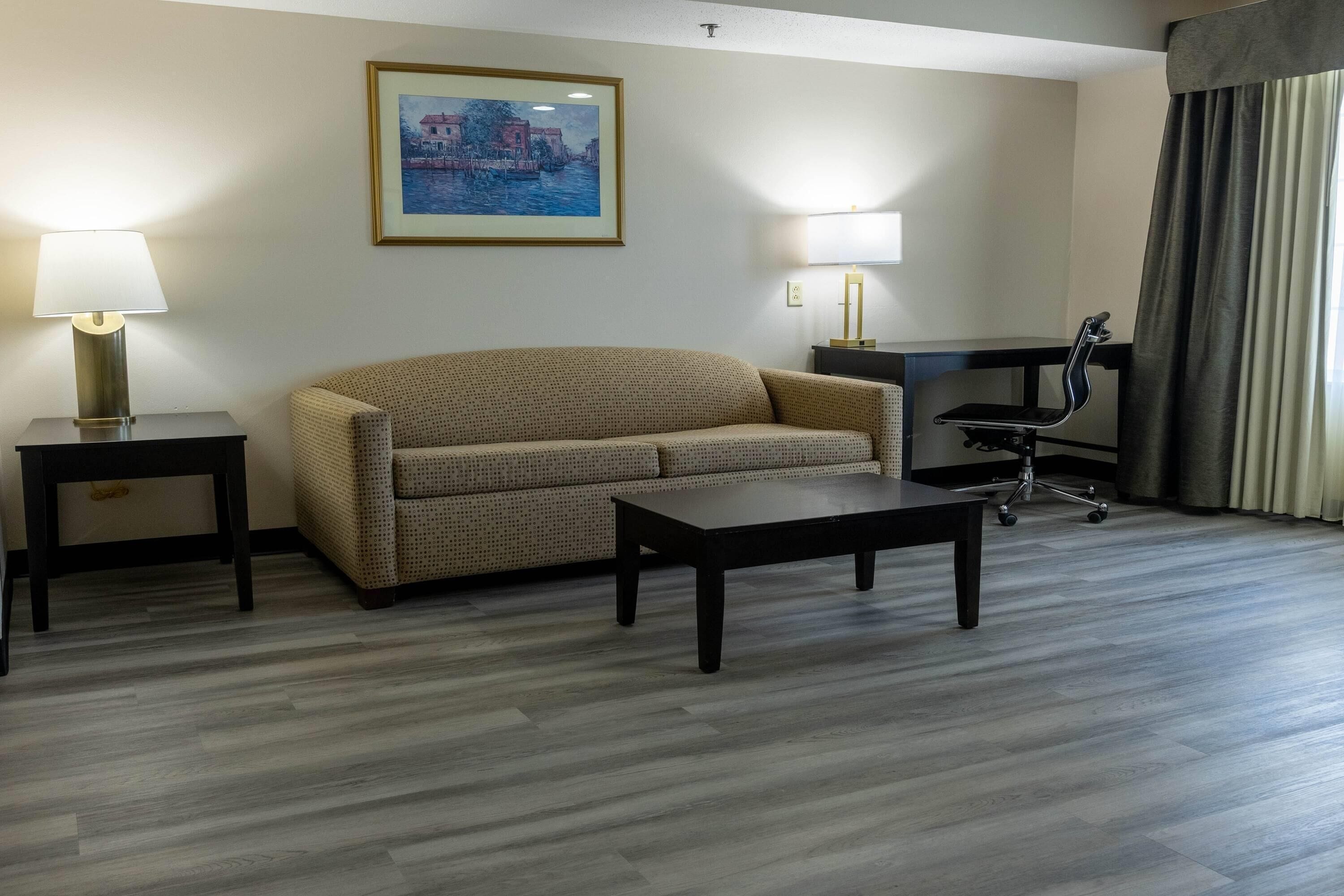 A hotel room with a sofa, two bedside tables, a desk with a chair, and a painting on the wall.