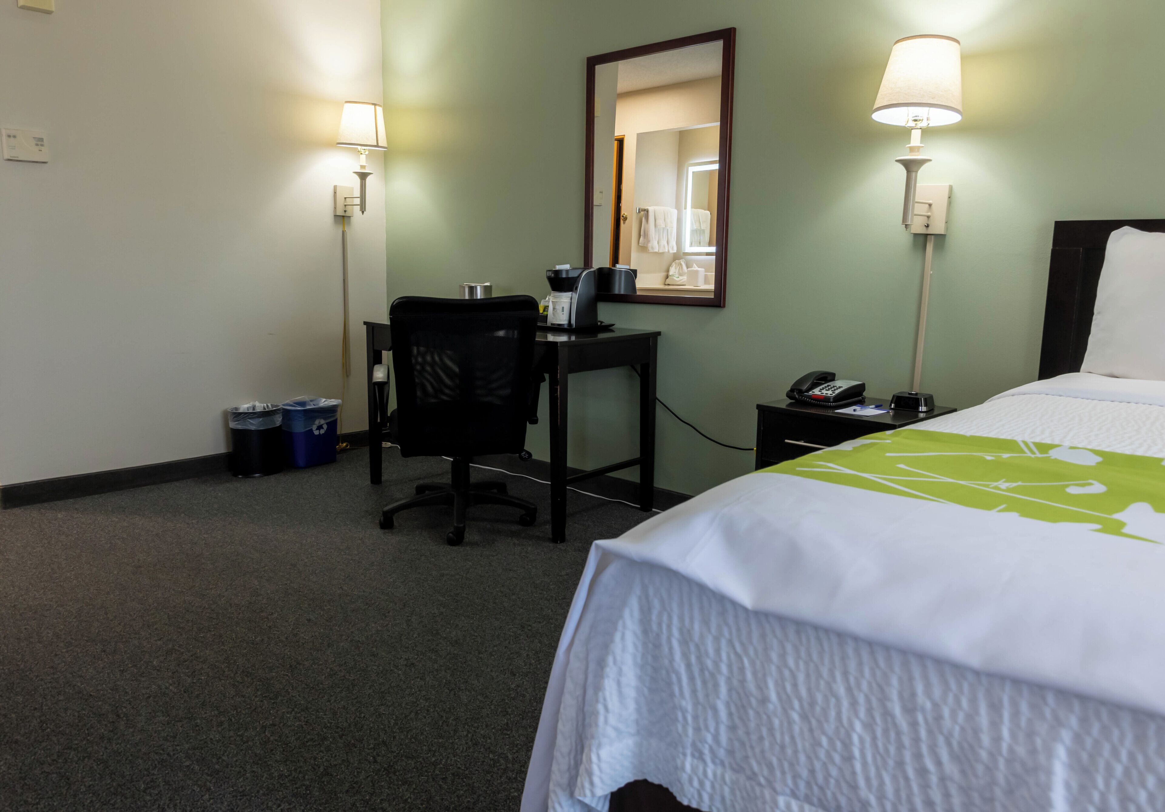 A hotel room with a bed, a desk, a chair, a lamp, a phone, and a trash bin.