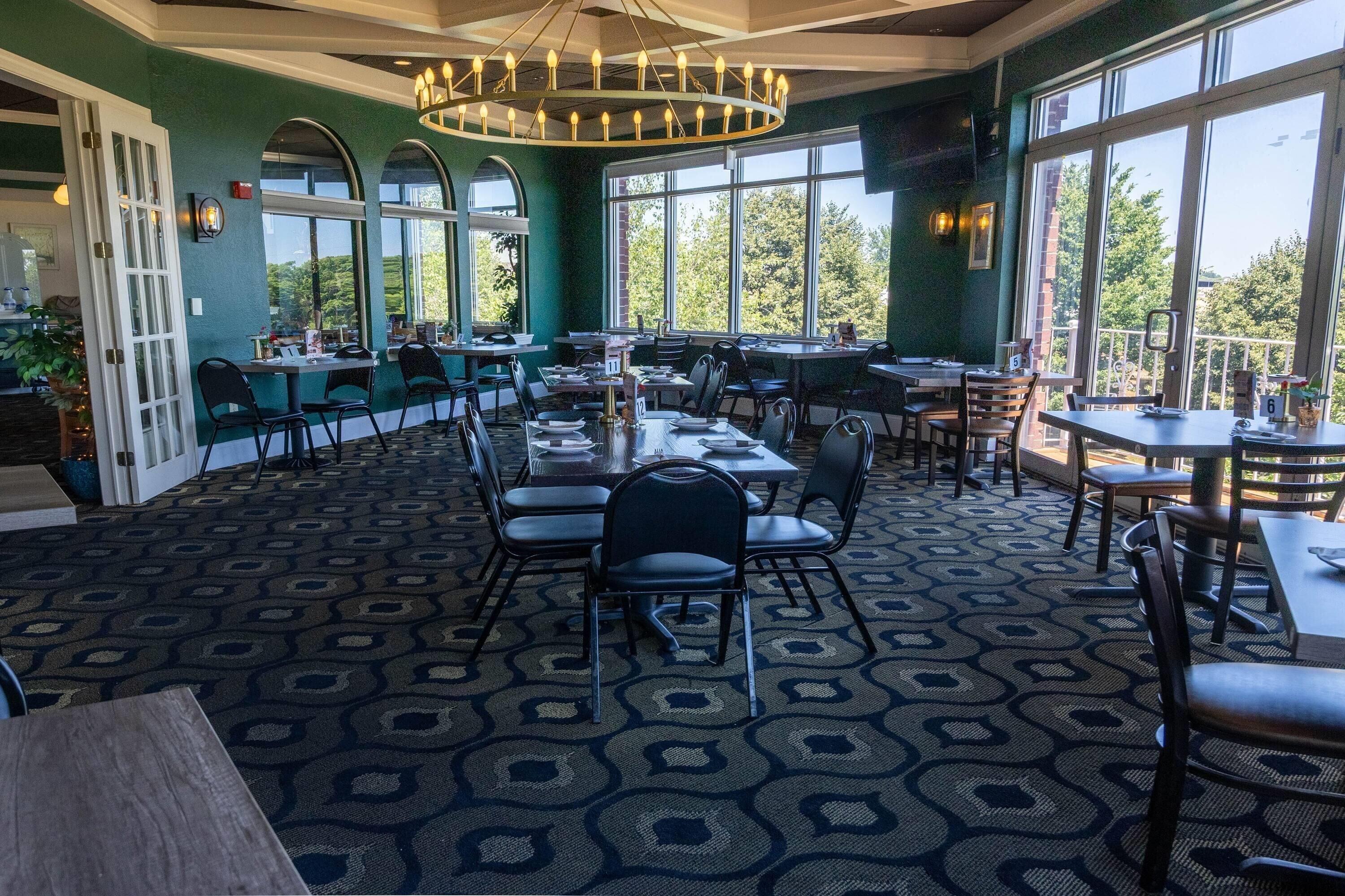 A spacious dining area with multiple tables and chairs, large windows offering a view of the outdoors, and a patterned carpet.