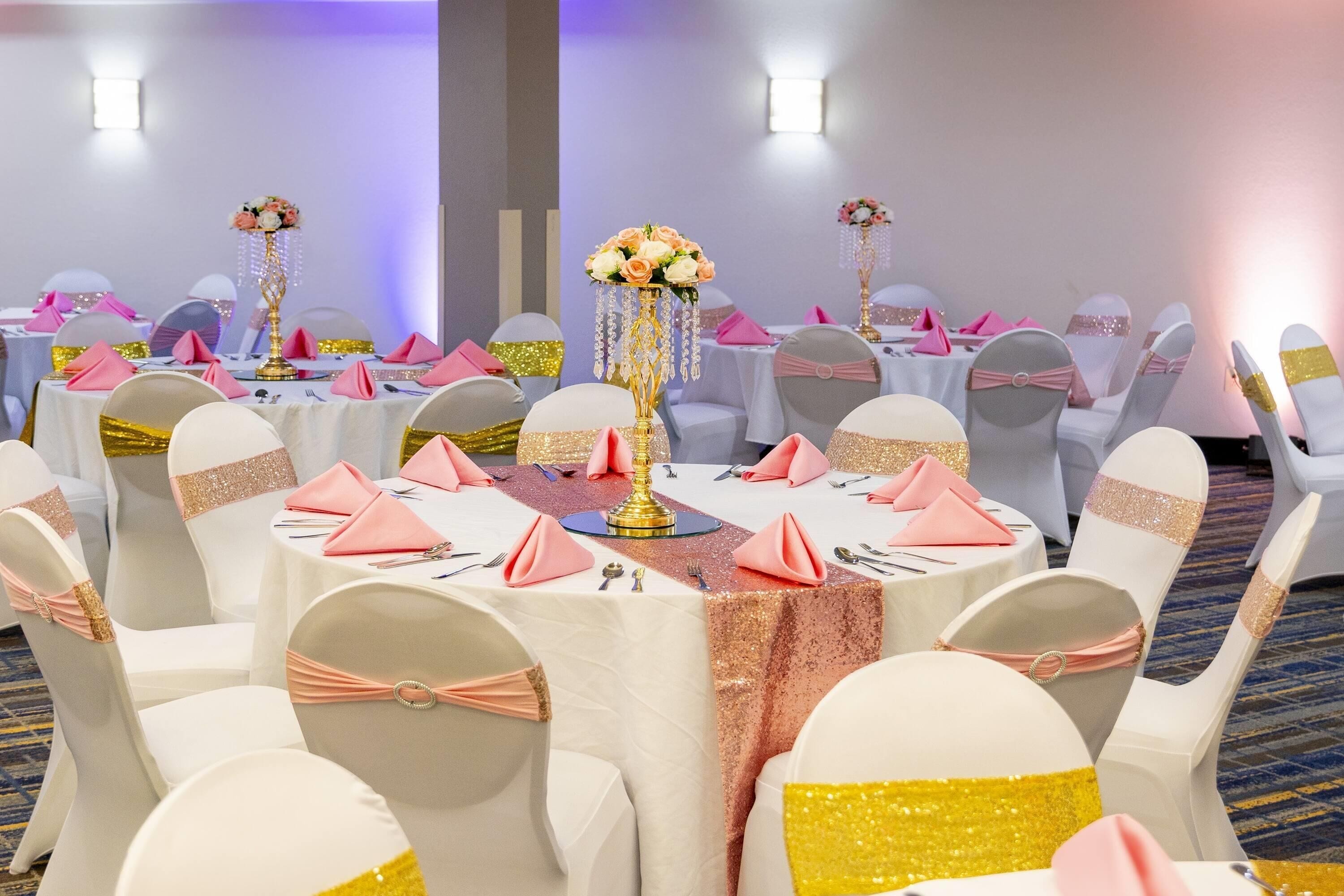 A banquet hall with round tables, white chairs with gold sashes, pink napkins, and floral centerpieces.