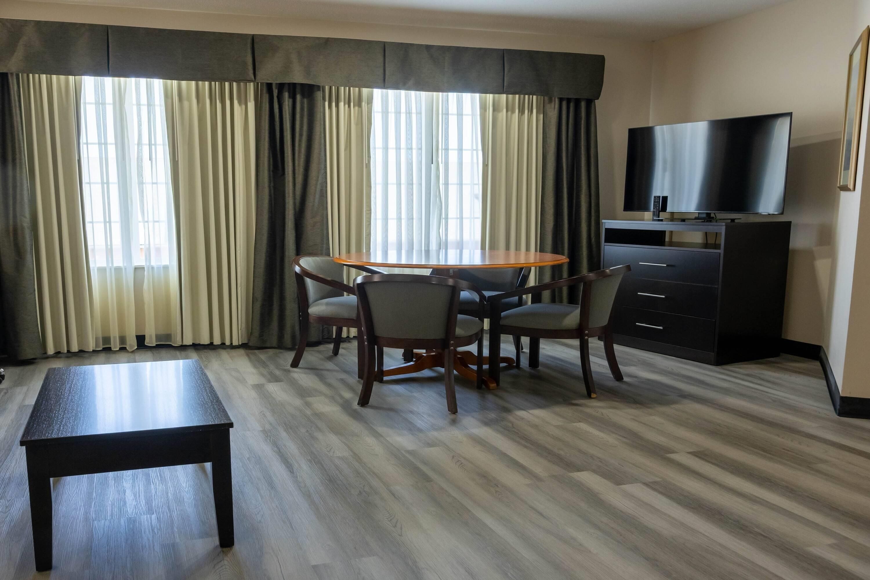 A hotel room with a dining table, chairs, a bench, a TV, and a cabinet.