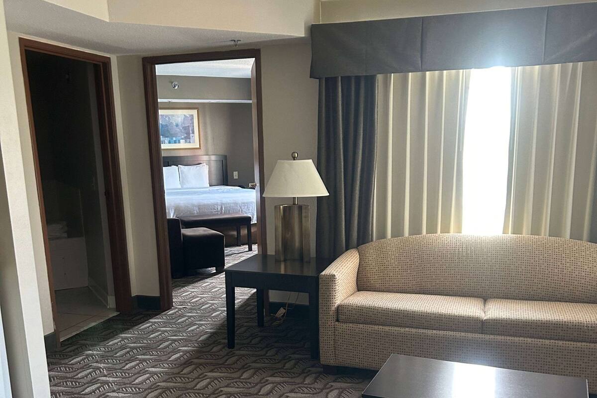 A hotel room with a bed, a sofa, a table, and a lamp. There is a window with curtains and a door leading to another room.
