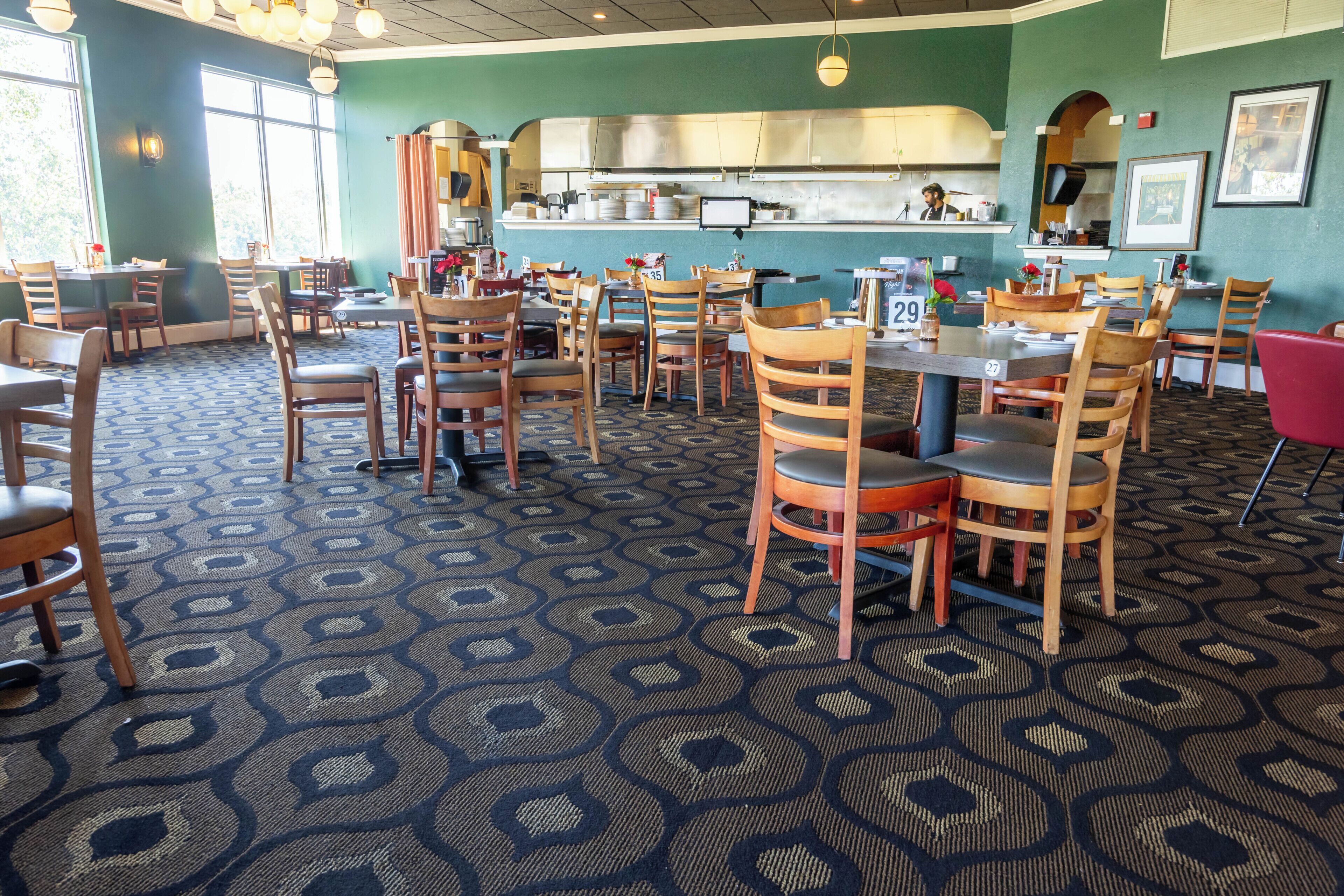 A restaurant interior with multiple tables and chairs, a kitchen in the background, and patterned carpet flooring.