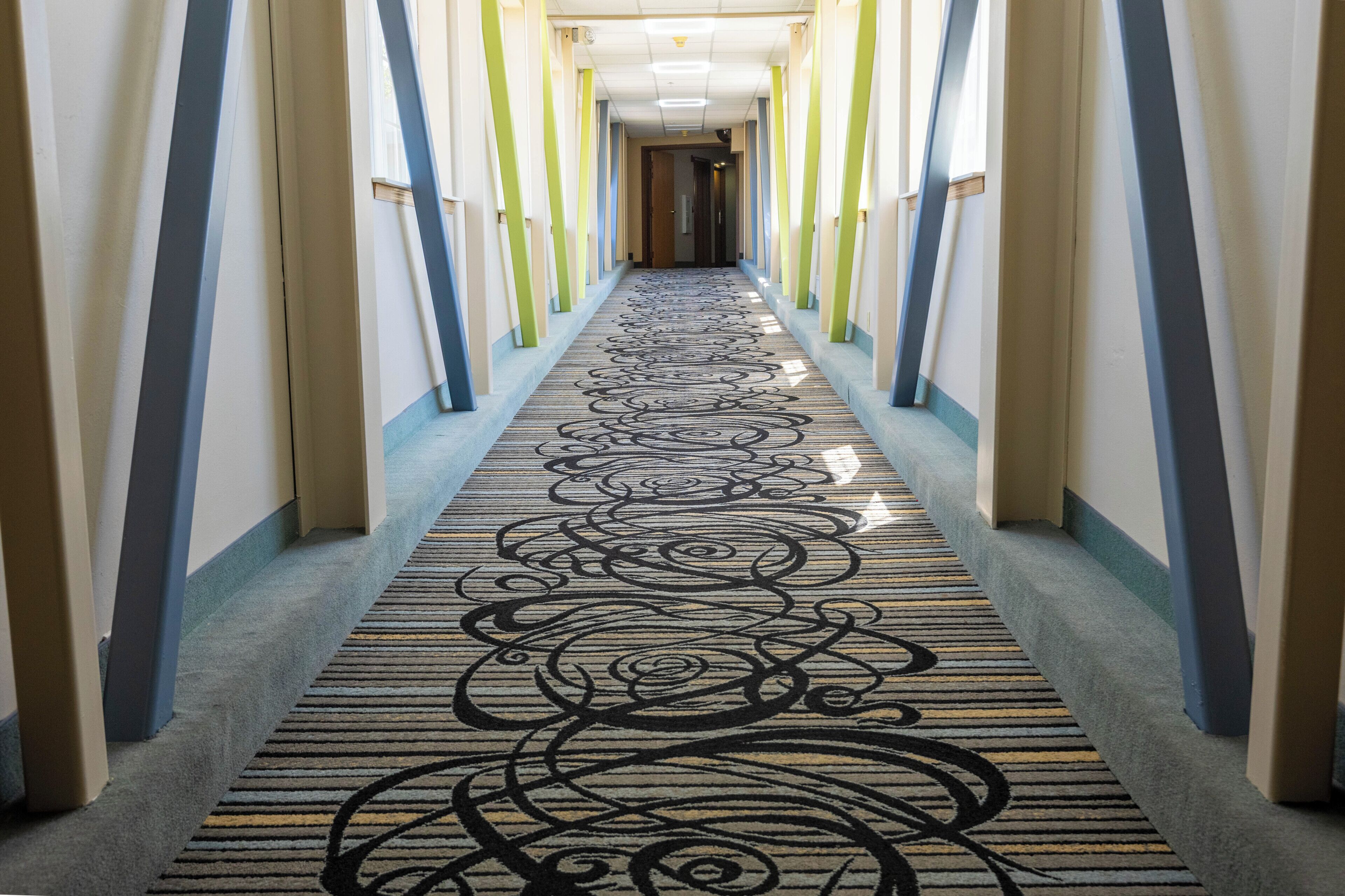 Un couloir d’hôtel avec un tapis à motifs, des colonnes verticales et une porte au fond.