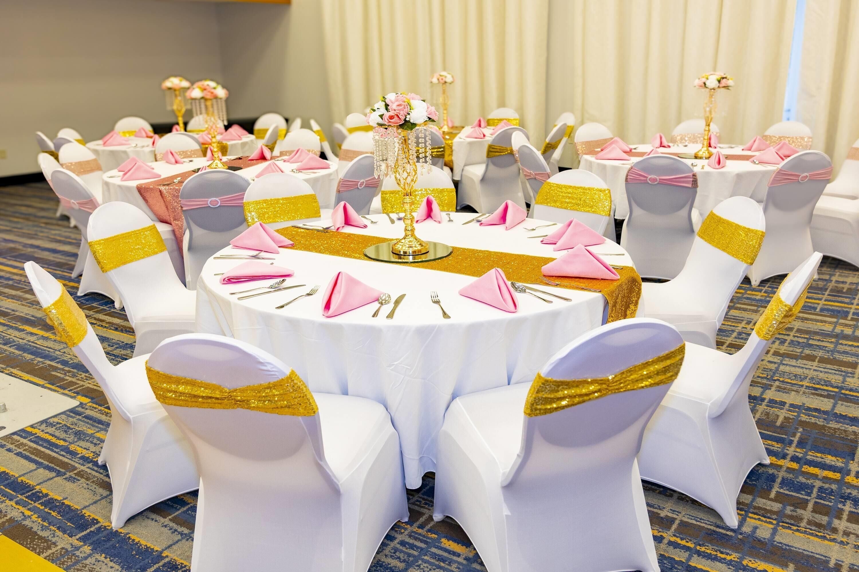 A round table with white tablecloths, gold sashes, and pink napkins.