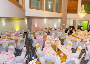 A banquet hall with round tables, white chairs with gold sashes, and a chandelier. - Wyndham Garden Madison Fitchburg (Madison)