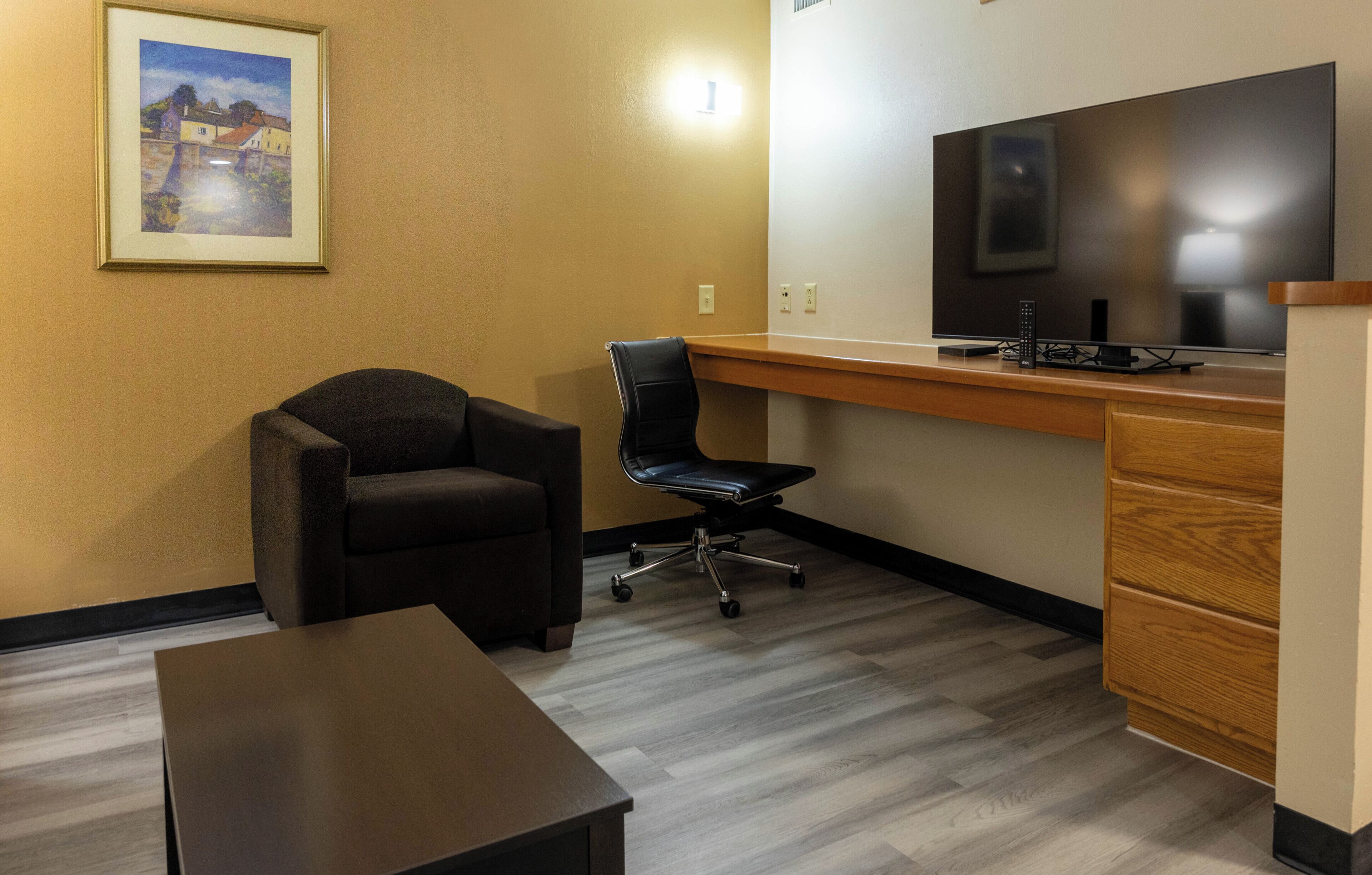 A hotel room with a desk, a chair, a television, and a painting on the wall.