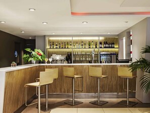 A modern bar area with a wooden counter, three bar stools, and a variety of bottles and glasses on shelves. - ibis Barcelona Cornella (Cornella de Llobregat)