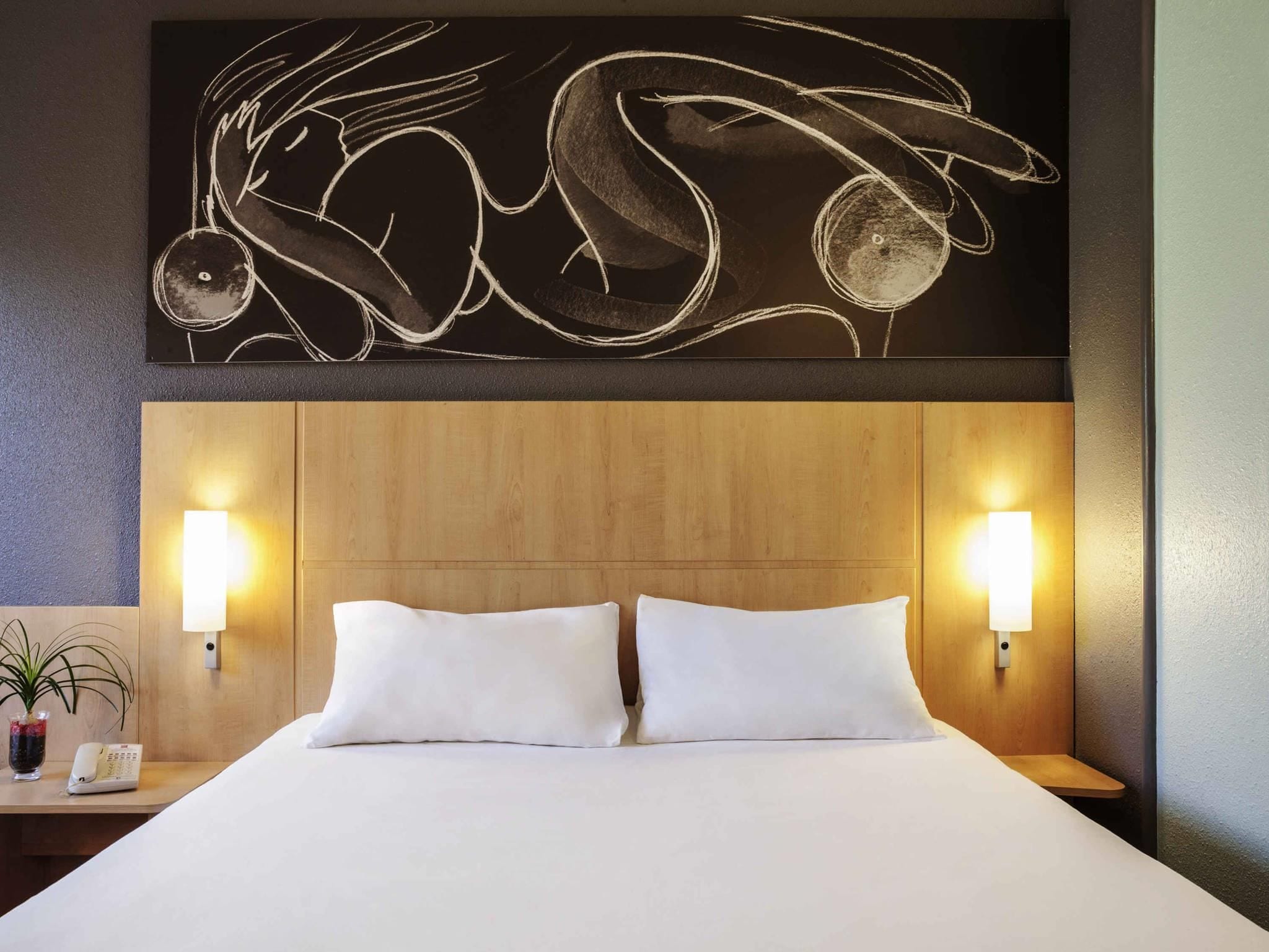 Une chambre d’hôtel avec une tête de lit en bois, deux oreillers blancs, une table de chevet avec une lampe et une œuvre d’art murale au-dessus du lit.