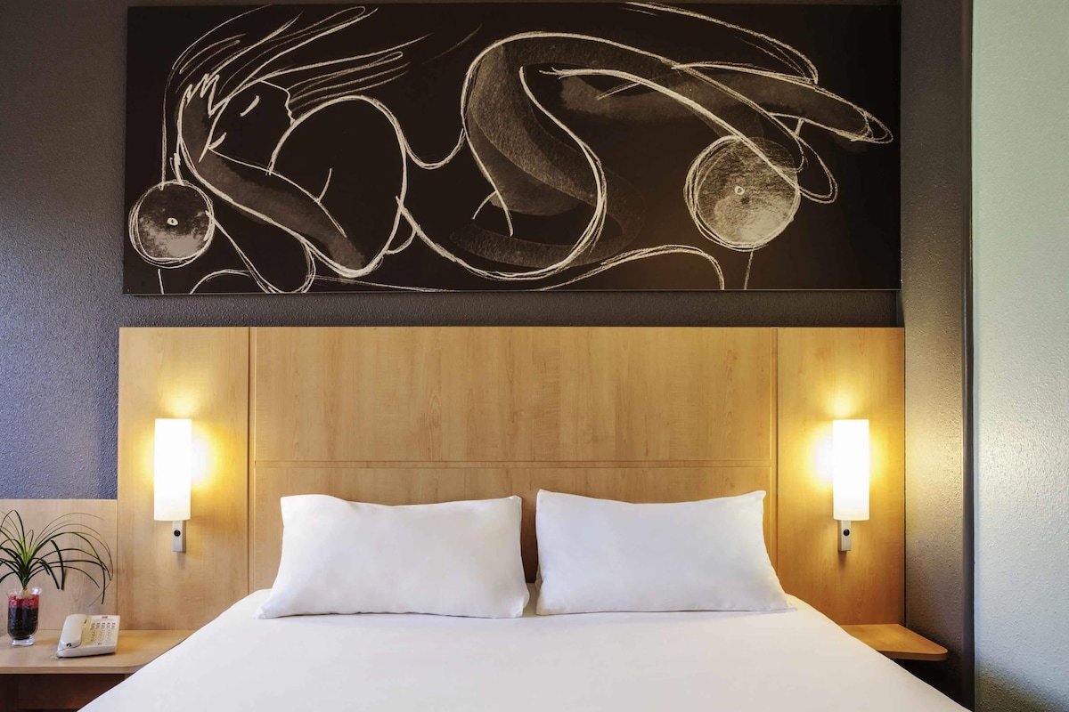 A hotel room with a wooden headboard, two white pillows, a bedside table with a lamp, and a wall art piece above the bed.