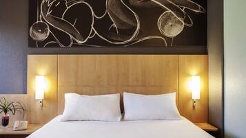 Une chambre d’hôtel avec une tête de lit en bois, deux oreillers blancs, une table de chevet avec une lampe et une œuvre d’art murale au-dessus du lit.