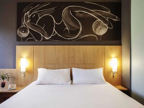 A hotel room with a wooden headboard, two white pillows, a bedside table with a lamp, and a wall art piece above the bed. - ibis Barcelona Cornella (Cornella de Llobregat)
