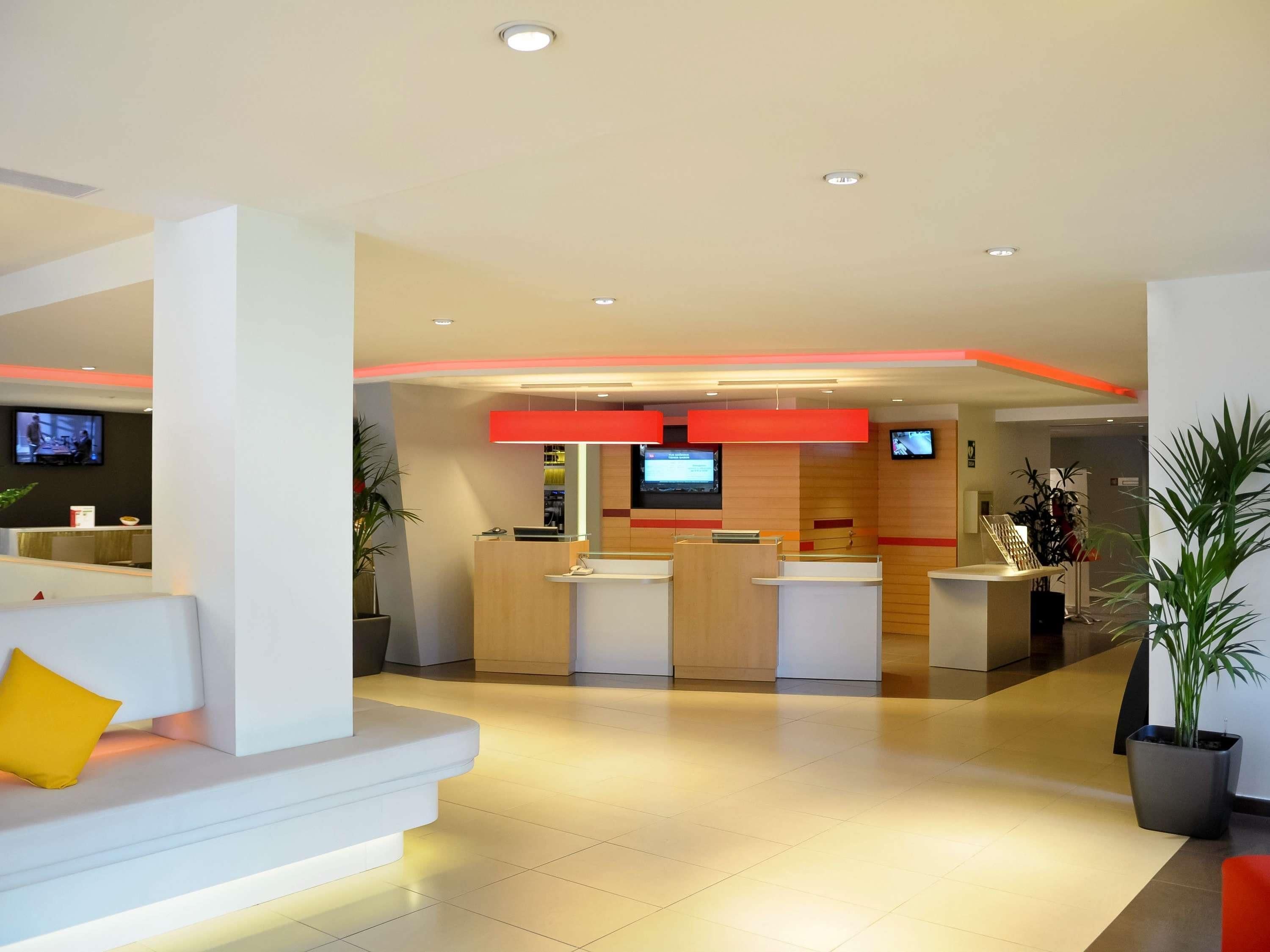 A modern hotel lobby with a reception desk, seating area, and a TV screen.