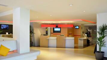 A modern hotel lobby with a reception desk, seating area, and a TV screen.