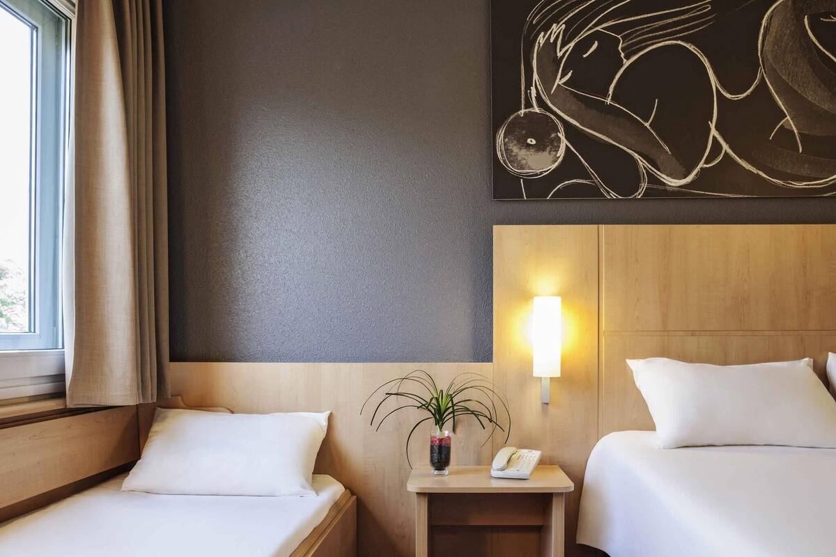 A hotel room with two beds, a bedside table with a lamp, a vase with a plant, and a wall-mounted artwork.