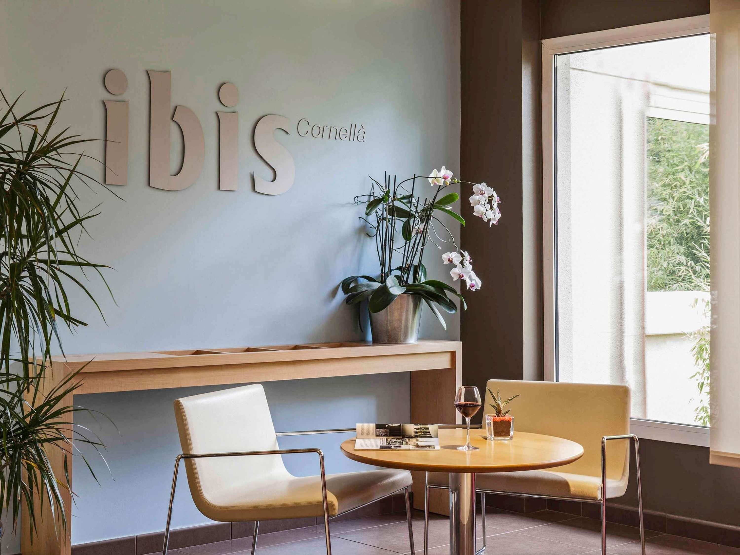 Un interior moderno con un letrero en la pared que dice 'ibis Cornella', una mesa redonda de madera con sillas, una planta en maceta y una ventana con vistas.