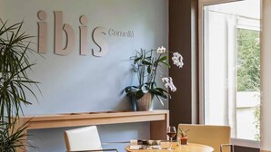 A modern interior with a wall-mounted sign reading 'ibis Cornella,' a round wooden table with chairs, a potted plant, and a window with a view. - ibis Barcelona Cornella (Cornella de Llobregat)