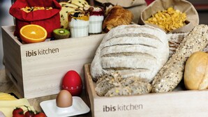 A wooden box labeled "ibis kitchen" containing various breads, a croissant, and a red egg with a timer. - ibis Barcelona Cornella (Cornella de Llobregat)