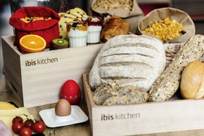 A wooden box labeled "ibis kitchen" containing various breads, a croissant, and a red egg with a timer. - ibis Barcelona Cornella (Cornella de Llobregat)