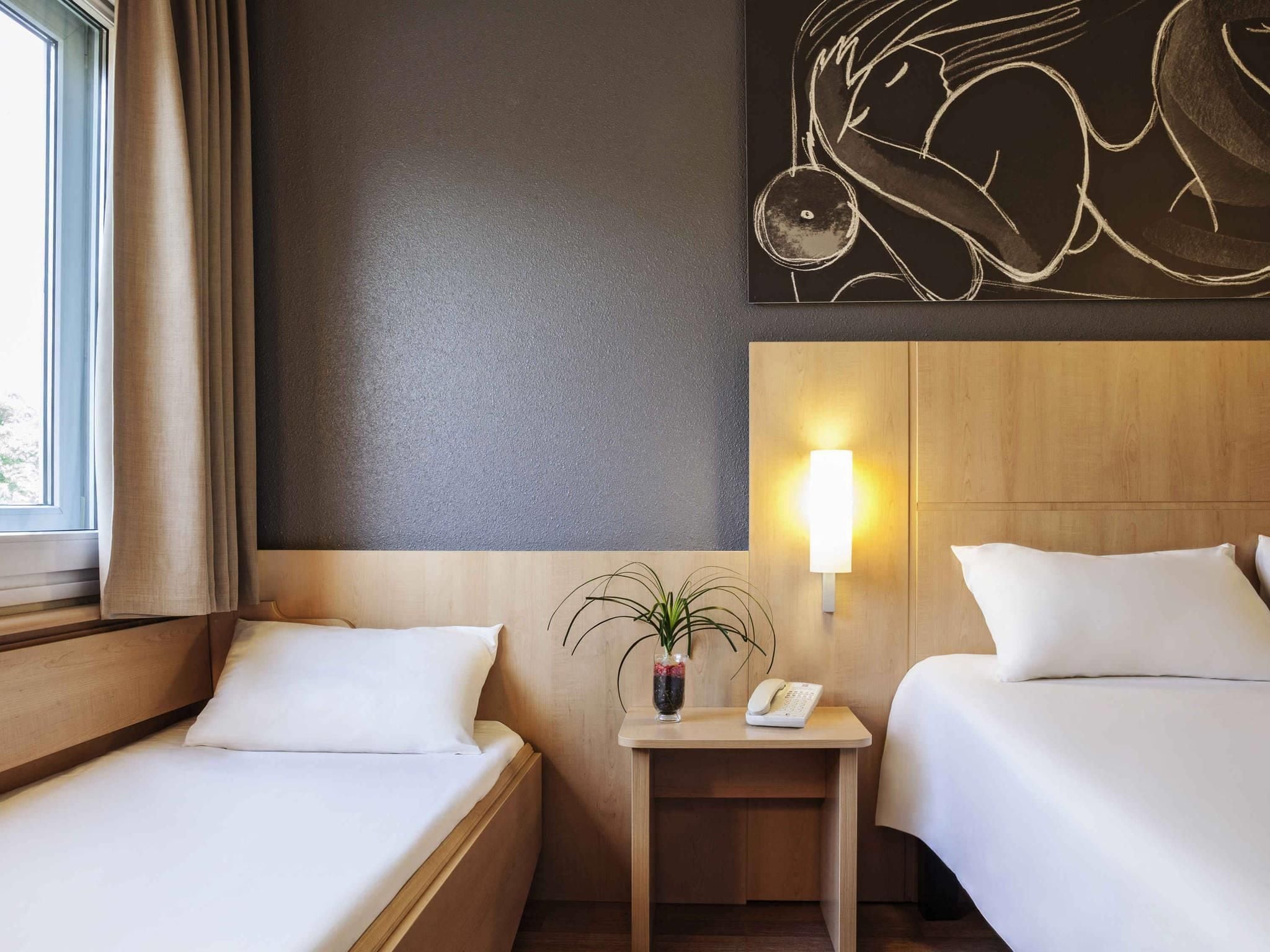 A hotel room with two beds, a bedside table with a lamp, a vase with a plant, and a wall-mounted artwork.
