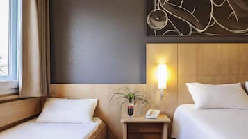 A hotel room with two beds, a bedside table with a lamp, a vase with a plant, and a wall-mounted artwork.