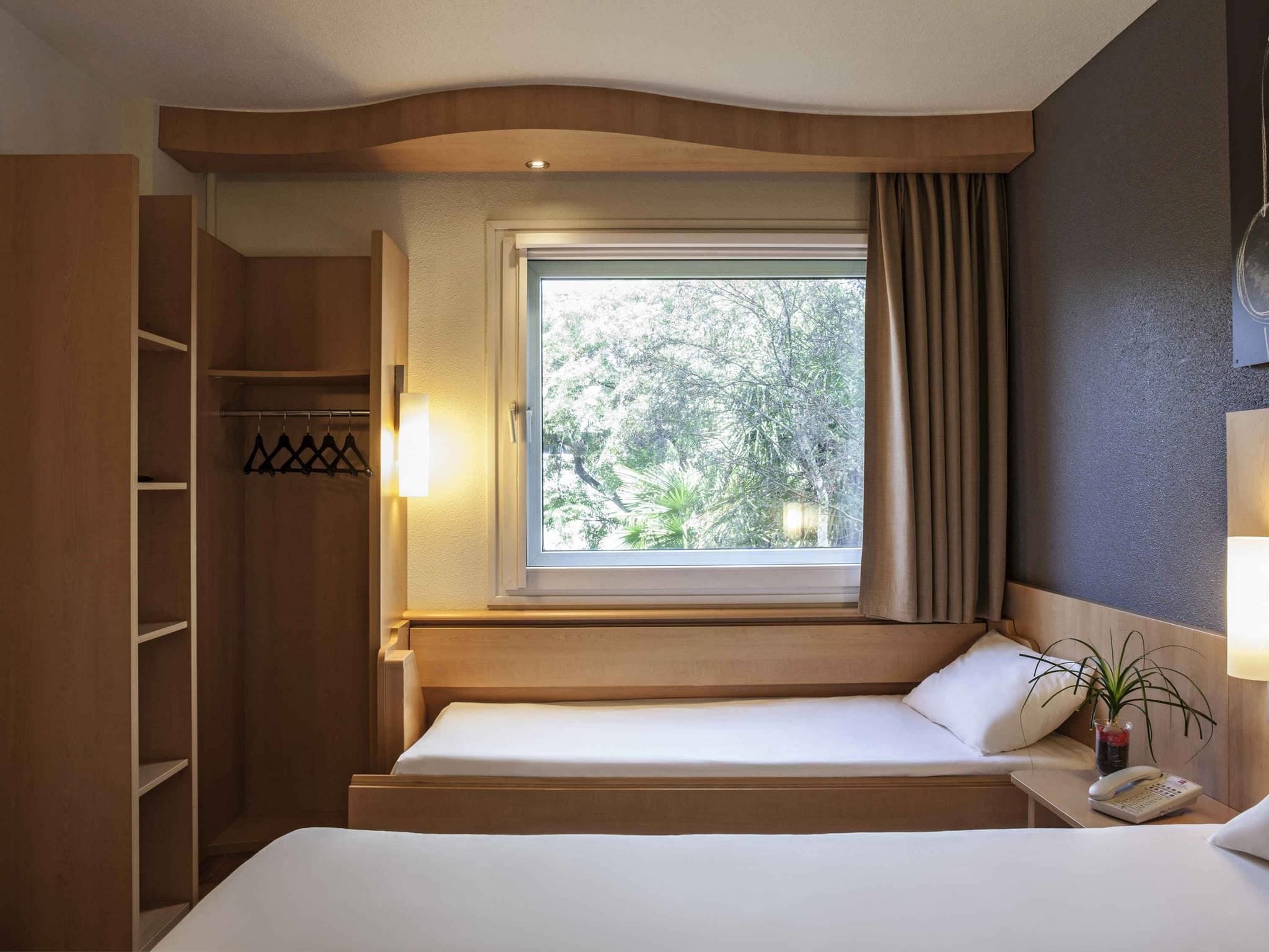 Une chambre d’hôtel avec un lit, une fenêtre avec des rideaux, une table de chevet avec une lampe, un téléphone et une plante.