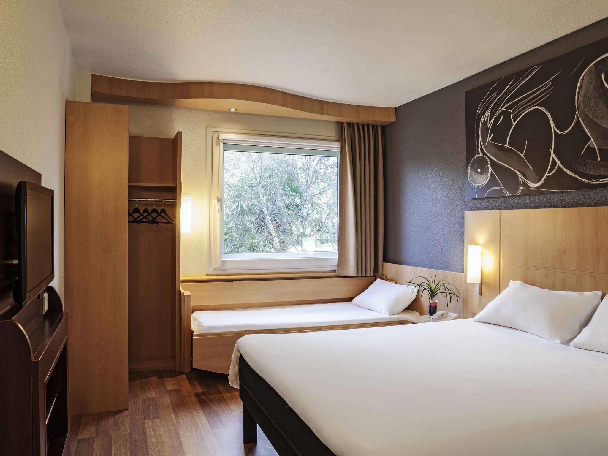 Una habitación de hotel con cama, mesita de noche, lámpara y ventana con cortinas.