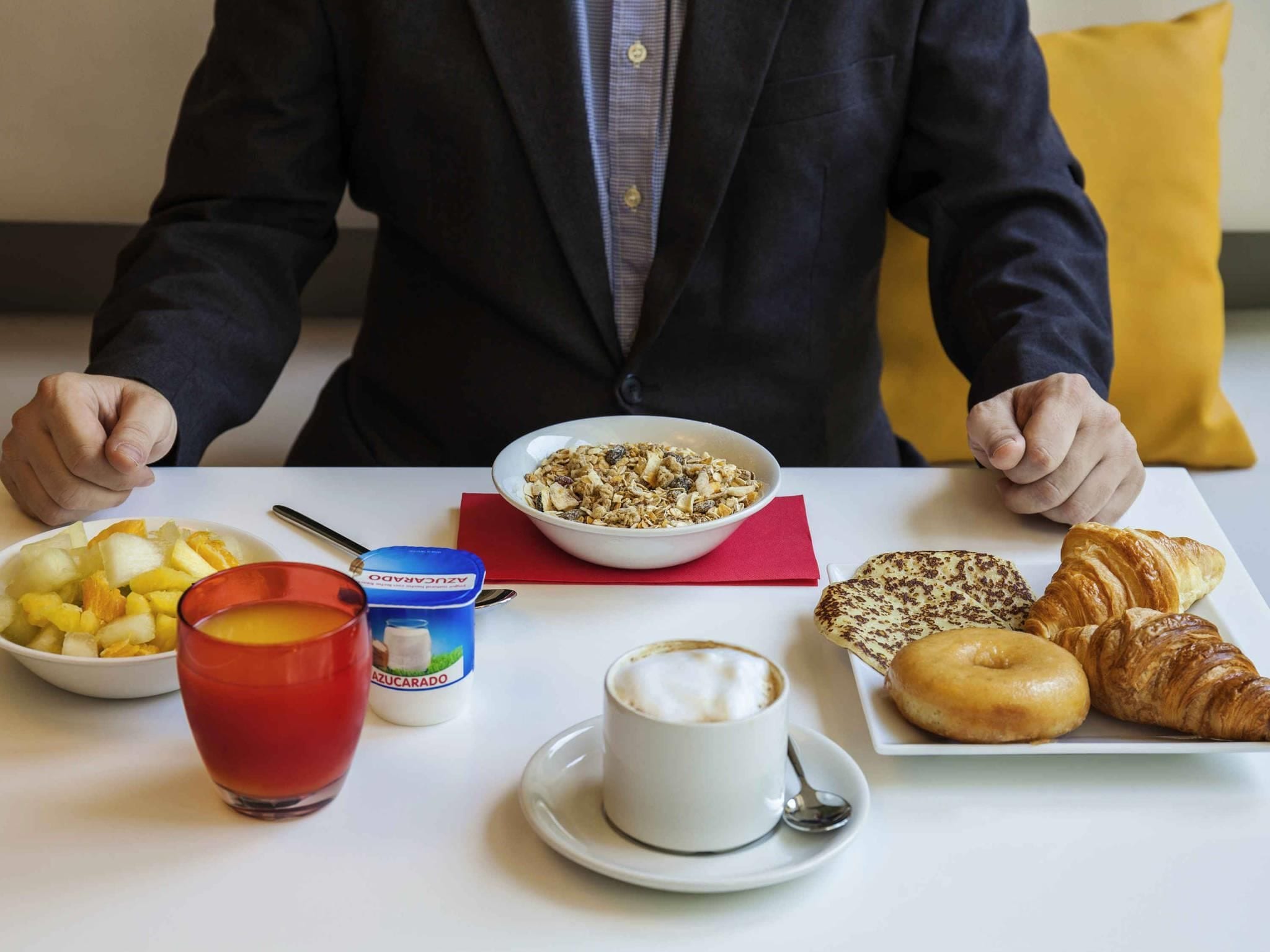 Aamiaispöydällä on kulho täynnä muroja, lautanen täynnä hedelmiä, lasi appelsiinimehua, maitotölkki, croissant, donitsi ja kuppi kahvia.