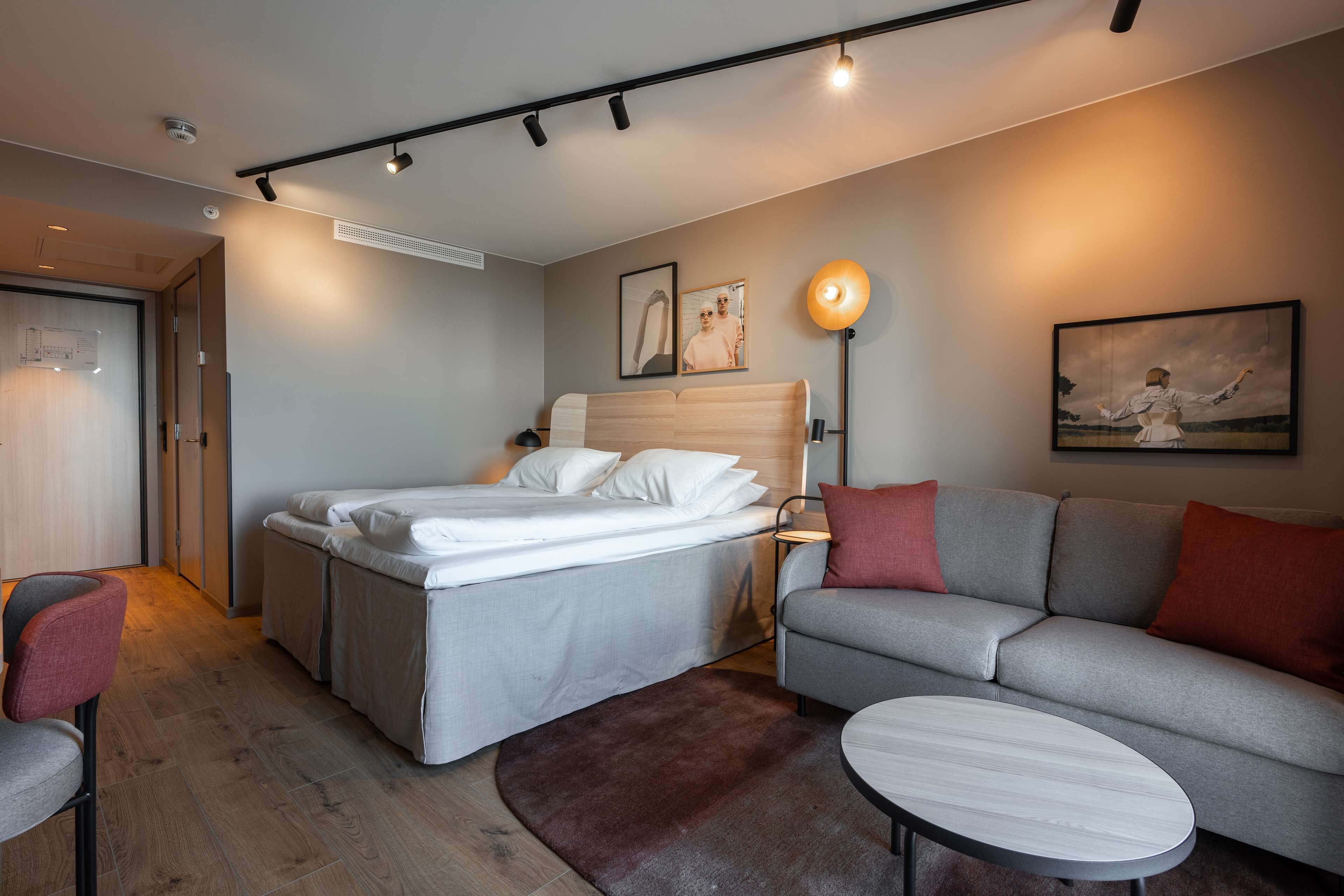 A modern hotel room with a bed, a grey sofa, a small round table, and a wall-mounted lamp.