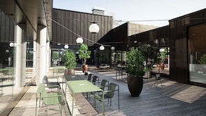 Un patio al aire libre con mesas, sillas, plantas en macetas y luces colgantes.