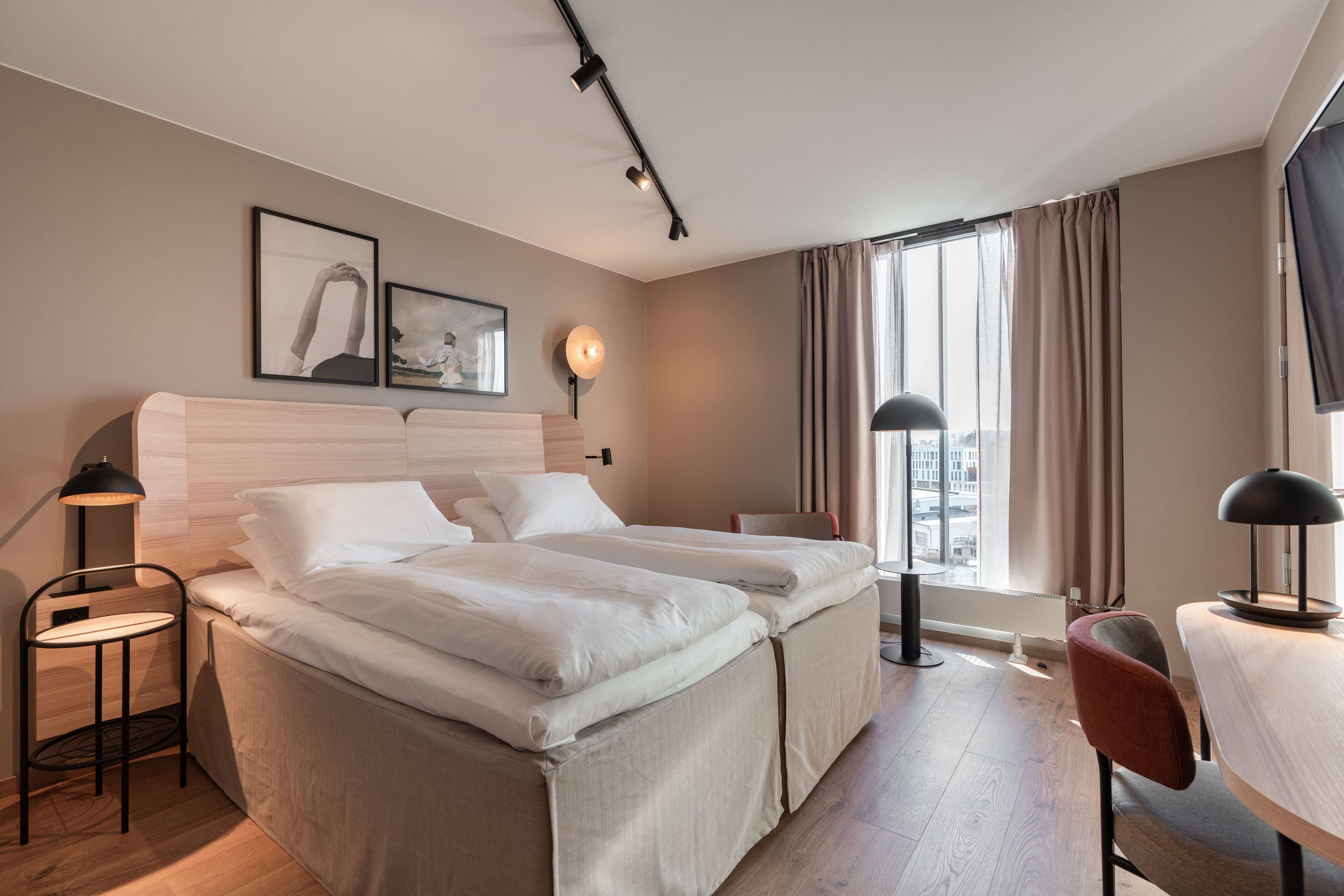 A hotel room with two beds, a desk, a chair, a lamp, a picture on the wall, and a window with curtains.