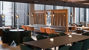 Un restaurante moderno con diversas opciones de asientos, incluyendo mesas y una zona con sofás, iluminación colgante y amplias ventanas.
