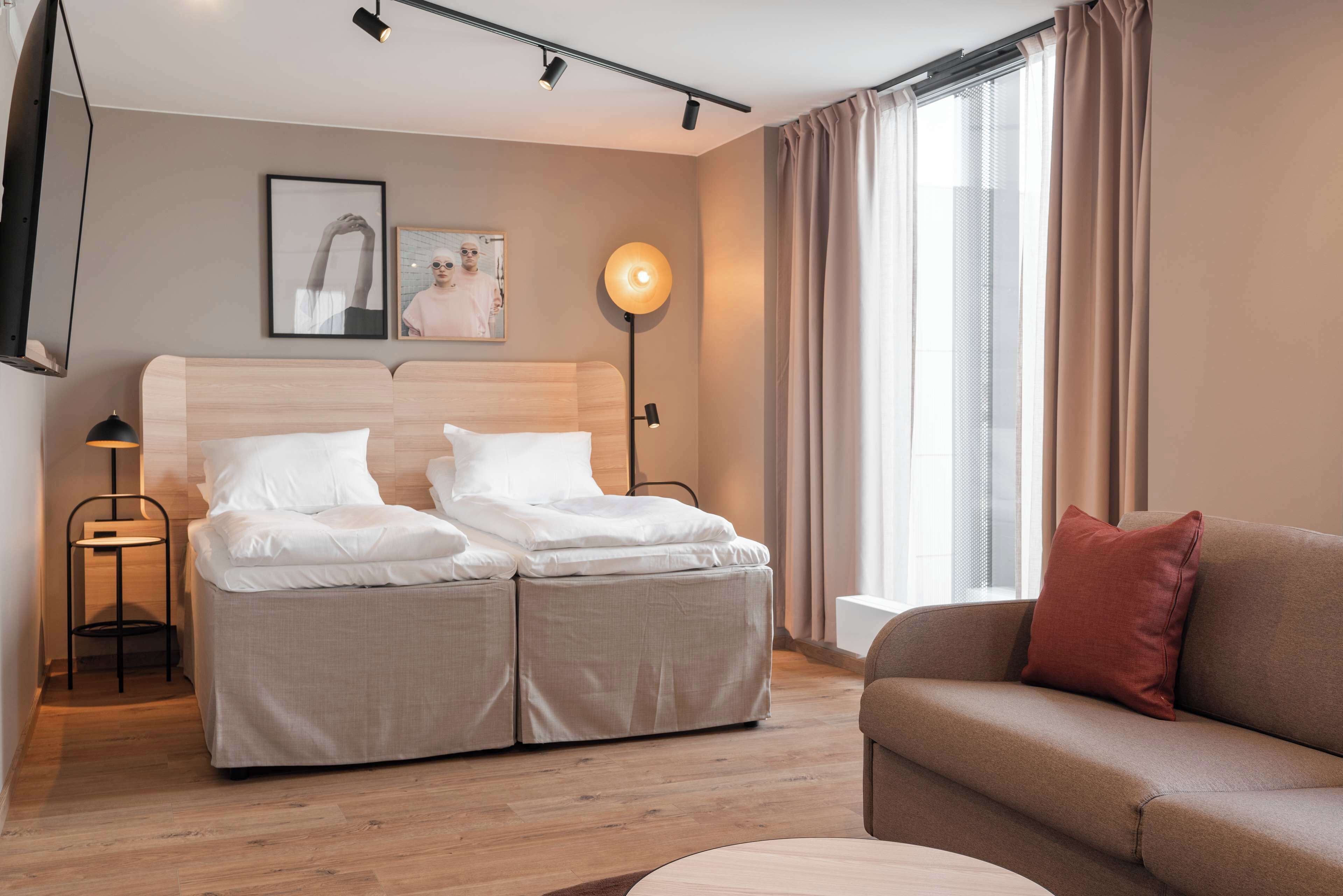 Une chambre d’hôtel moderne avec un lit double, un canapé, une lampe sur pied et une grande fenêtre avec des rideaux.