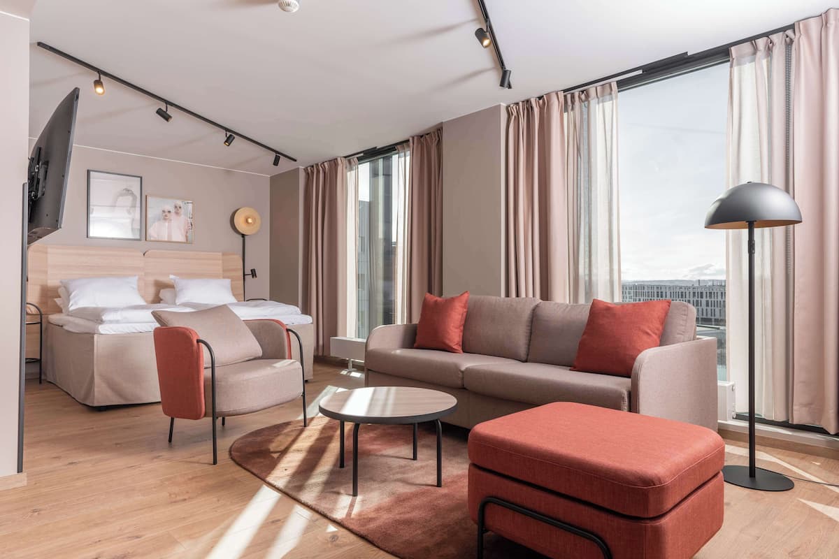 A modern hotel room with a sofa, armchair, and ottoman, a bed, a small table, and a floor lamp.