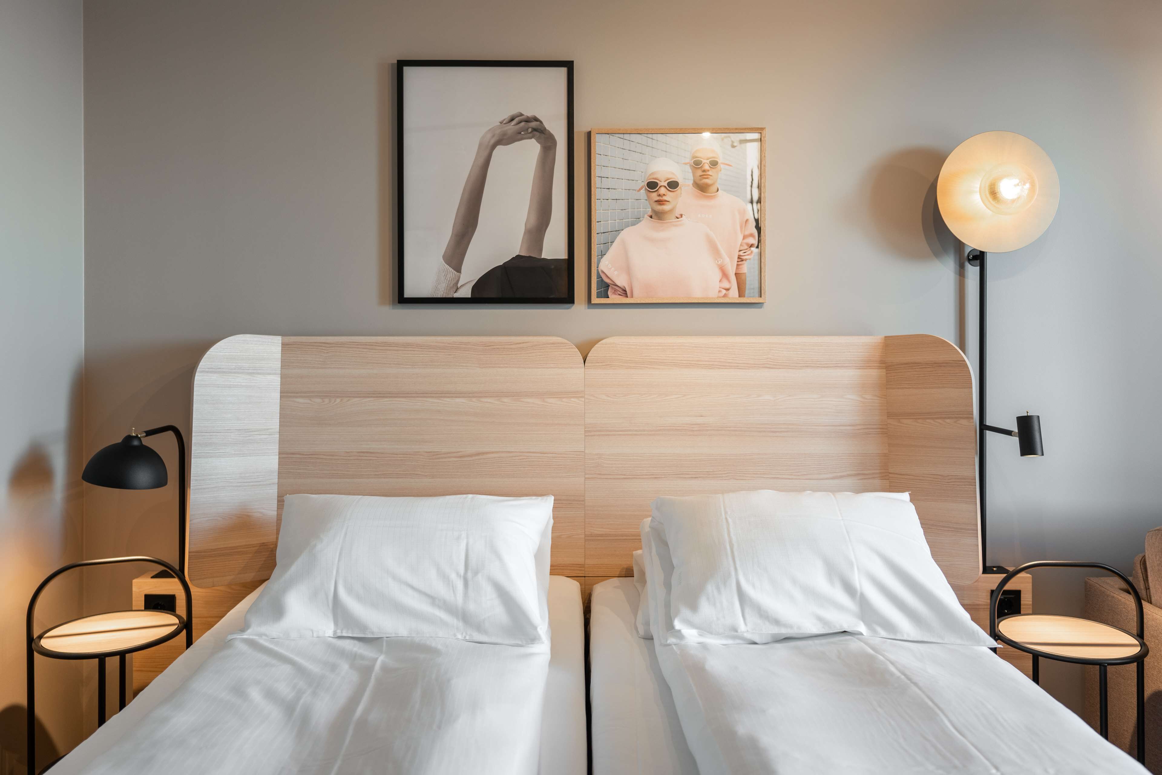 Un lit double avec du linge de lit blanc, une tête de lit en bois, deux tableaux encadrés au mur, une lampe de table ronde et une petite table ronde.