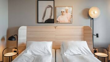 A double bed with white linens, a wooden headboard, two framed pictures on the wall, a round table lamp, and a small round table.