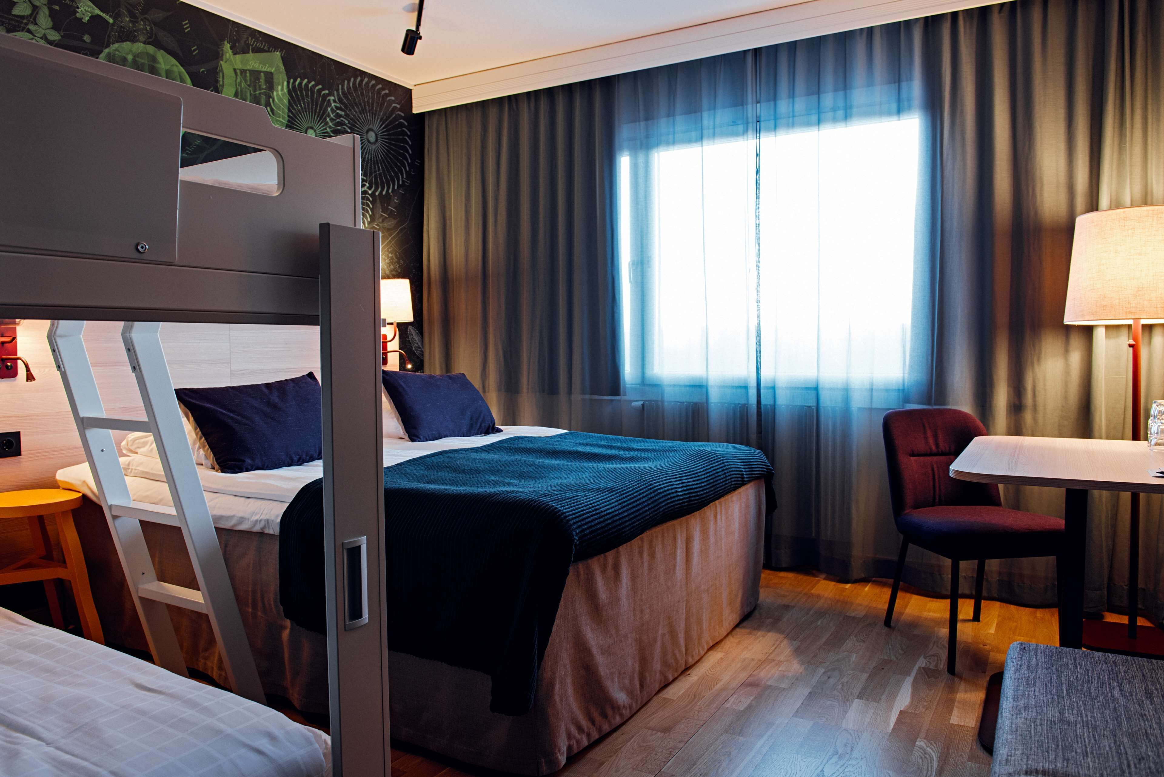 A hotel room with a bunk bed, a desk, a chair, a lamp, and a large window with curtains.