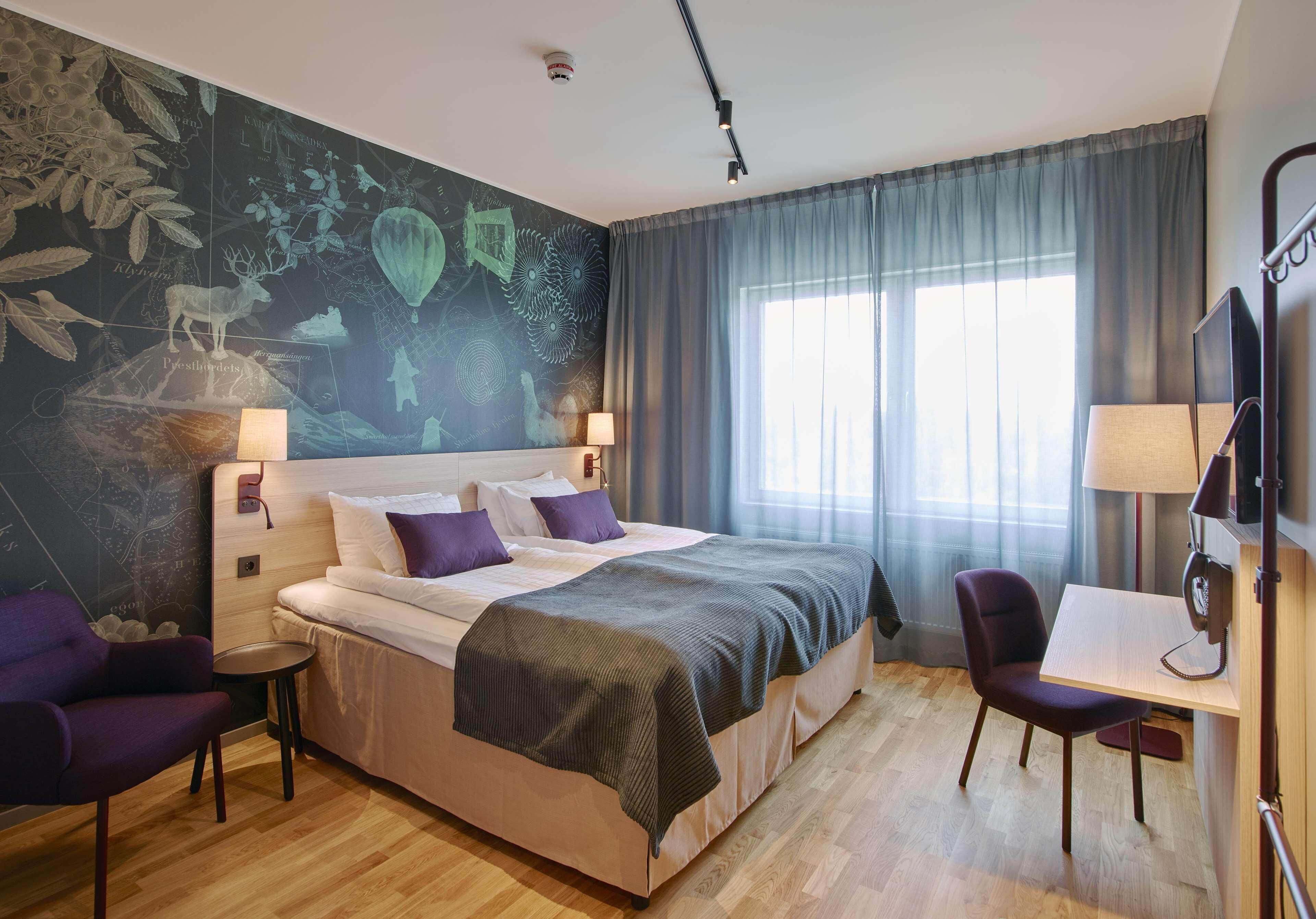 A modern hotel room with a large bed, a desk, a chair, and a mural on the wall.